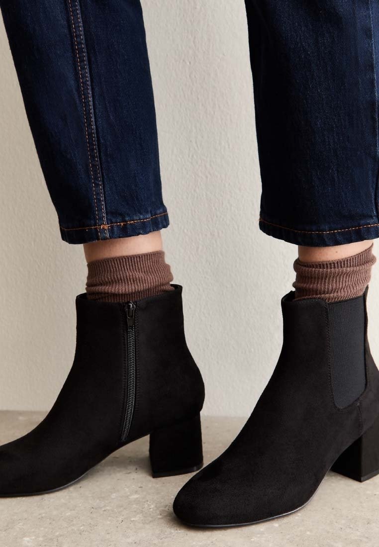 Bottes en daim noir montant à la cheville avec talons blocs, fermeture éclair latérale, portées avec des chaussettes marron et un jean bleu foncé sur un fond de mur clair.