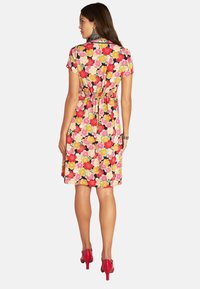 Blumenkleid mit einem lebhaften Muster in Rot, Pink und Gelb, kurzen Ärmeln, gerafftem Taillenbund und einer Länge bis über das Knie, kombiniert mit roten High Heels.