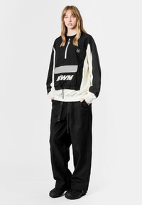 Suéter negro y crema con cremallera de cuarto, bolsillo frontal y detalle gráfico; combinado con pantalones anchos de textura negra y zuecos.