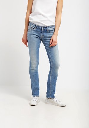 Persoon draagt lichtblauwe slim-fit jeans, wit mouwloos shirt en witte sneakers, staand tegen een effen witte achtergrond.