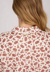 Chemise crème avec un motif paisley rouge détaillé, un col, et un léger pli à l'arrière.