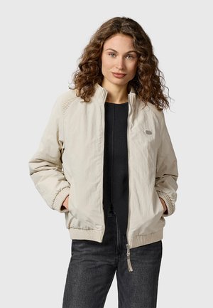 Kobieta o kręconych włosach, ubrana w jasnobeżową kurtkę zapinaną na suwak na czarną bluzkę i ciemne dżinsy, stoi z rękami w kieszeniach kurtki.