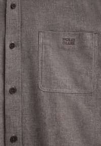 Brązowa koszula na guziki wykonana z teksturowanego materiału, z kieszenią na piersi ozdobioną logo "POLO CLUB" oraz ciemnymi guzikami na przodzie.