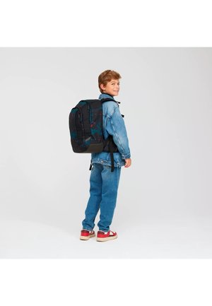 Ragazzo con giacca di jeans e jeans indossa un grande zaino nero con motivi blu e rossi, si gira indietro sorridendo.