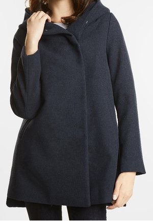 Kobieta ubrana w ciemnogranatowy wełniany płaszcz z kapturem i długimi rękawami, trzymająca kołnierz jedną ręką, na jednolitym tle.