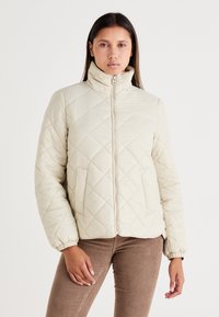 Lysebeige quiltet pufferjakke med høj krave, lynlås foran og elastiske manchetter. Har diagonale syninger og sidelommer.