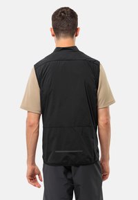 Gilet nero senza maniche con una texture leggera e liscia; presenta un colletto e un sottile dettaglio orizzontale sul retro. Abbinato a una camicia beige.