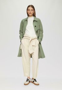 Groene trenchcoat met zwarte knopen, witte gebreide trui om de taille geknoopt, beige broeken en zwart-witte sneakers met beige zolen.