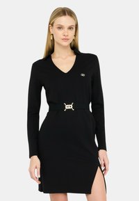 Abito nero a maniche lunghe con scollatura a V, vita sfrangiata e un accento in oro su una cintura nera. Presenta uno spacco laterale e una texture di tessuto morbido.