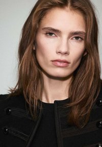 Zbliżenie portretu modelki z długimi, prostymi brązowymi włosami. Ma na sobie czarną kurtkę w poziome paski z dużymi czarnymi guzikami, wykonana z teksturowanej tkaniny.