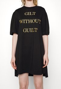 Černé tričkové šaty s krátkým rukávem, s výrazným zlatým textem "GILT WITHOUT GUILT" na přední straně, s postranními rozparky na spodním lemu.