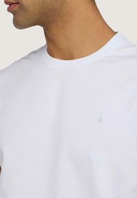 Camiseta de algodón blanca con cuello redondo clásico, que presenta un sutil logo gris claro en el pecho y una textura suave.