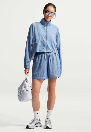 Vrouw draagt een lichtblauw jasje met rits en bijpassende korte broek, houdt een witte zachte tas vast, met zilverkleurige sneakers en sportieve zonnebril.