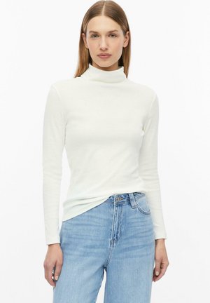 Frau mit glattem hellbraunem Haar, die ein weißes langärmeliges Rollkragenoberteil und hellblaue hoch taillierte Jeans trägt, steht vor weißem Hintergrund.