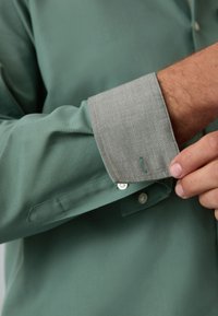 Camicia verde con bottoni, colletto e polsini grigio chiaro. Il polsino presenta un tessuto a contrasto e una piccola etichetta. Tessuto morbido con vestibilità sartoriale.