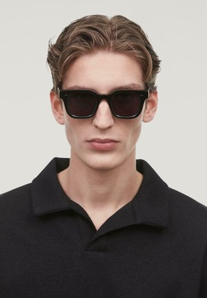 Lunettes de soleil noires avec des montures épaisses, des verres foncés et des détails subtils. Le modèle porte un haut noir à col avec une finition texturée.