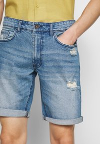 Pantalones cortos de mezclilla en azul claro con un lavado deslavado, dobladillos deshilachados y parches desgastados. Con cinco bolsillos y cierre de botón.