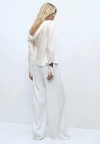 Halenka s dlouhým rukávem v barvě off-white, se splývavým zadním dílem a otevřenými manžetami, v kombinaci s volnými bílými kalhotami širokého střihu. Hladký povrch, minimalistický design.