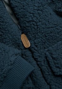 Donkerblauwe fleecejas met gestructureerde stof, geribbelde manchetten en een rits met een bruine leren trekker met het label "newbie".