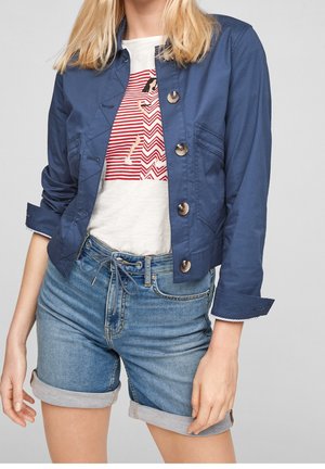 Frau trägt eine blaue Jacke mit Knöpfen über einem weißen T-Shirt mit rotem Hunde-Motiv und hochgekrempelte Jeansshorts.