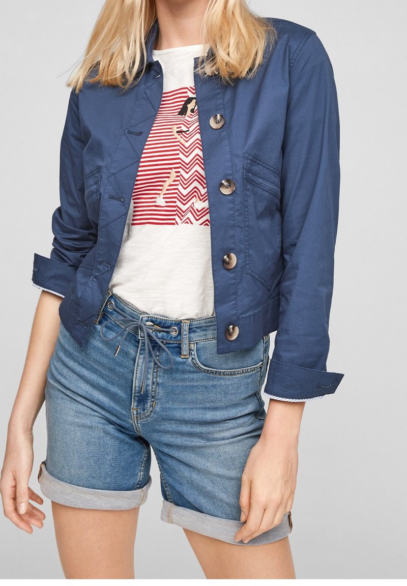 Femme portant une veste bleue à boutons sur un t-shirt blanc avec un motif de chien rouge et un short en jean retroussé.