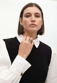 Schwarze gestrickte Weste, die über einem weißen Hemd mit püffen Ärmeln getragen wird; goldene Knopfakzente; große goldene Ohrringe; ein mehrfarbiger Ring.