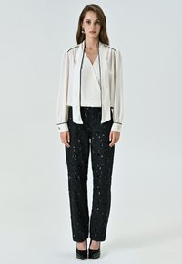 Camicia bianca con rifiniture nere, design a strati e maniche lunghe, abbinata a pantaloni neri con paillettes e tacchi neri. Look semplice ed elegante.