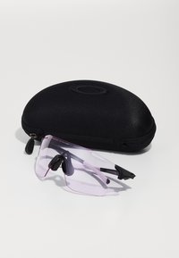 Sonnenbrille mit einer transparenten pinken Linse, schwarzen Bügeln und gummierten Akzenten neben einem schwarzen, mit Reißverschluss versehenen Schutzhülle.