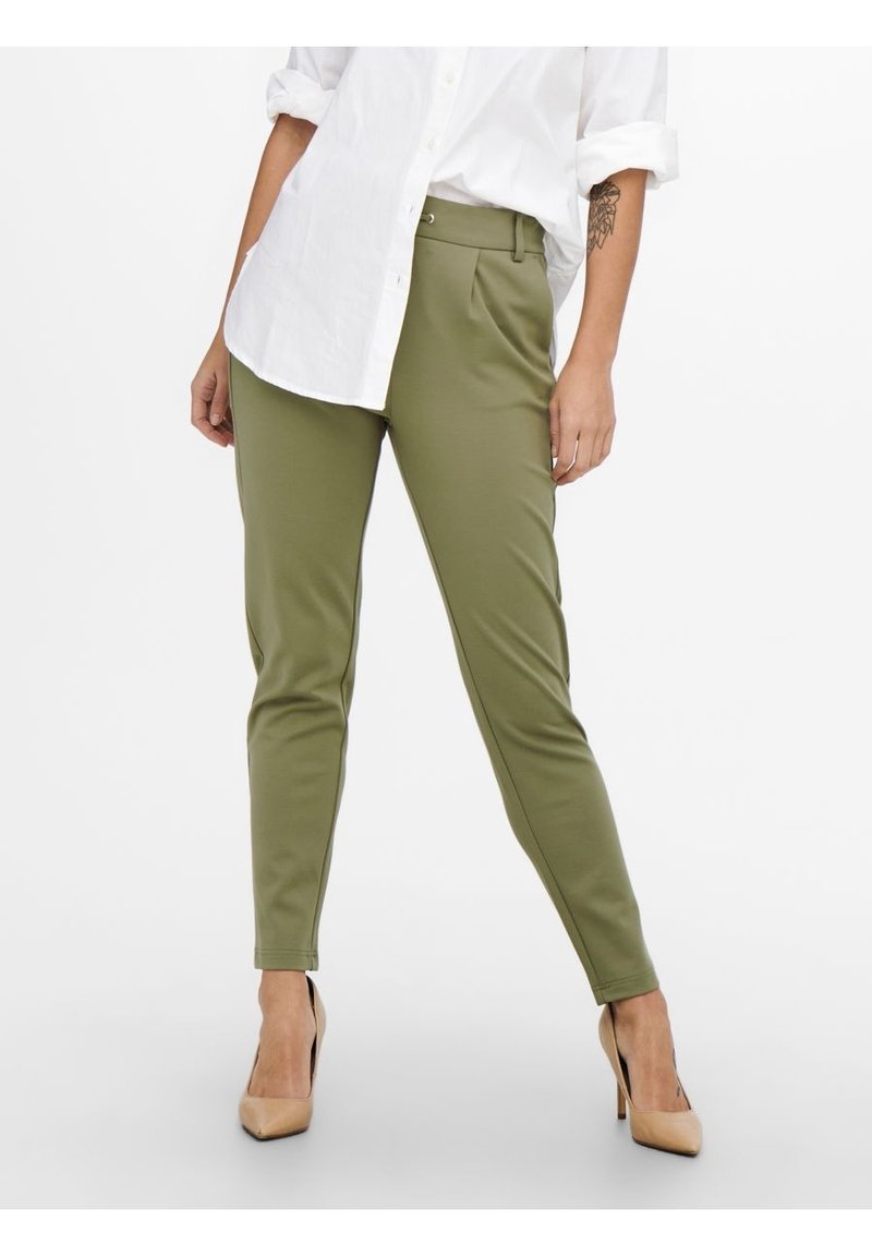 Femme portant un pantalon slim kaki avec une chemise blanche et des chaussures à talons beiges, debout devant un fond uni.