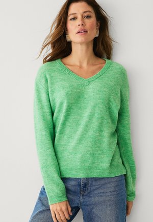 Kvinde iført en lysegrøn V-hals sweater og blå jeans, der står mod en ensartet lys baggrund, med guld øreringe i ringform.