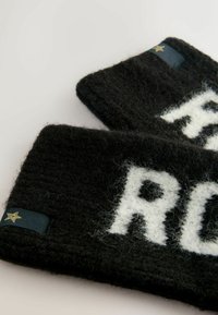 Czarna wełniana opaska na głowę z białym napisem. Zawiera małą złotą gwiazdkę jako akcent na metce. Fuzzy tekstura i prostokątny kształt.