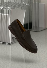 Mocassins en cuir marron foncé avec une texture lisse, des coutures sur les côtés et une silhouette élégante, posés sur un sol de couleur claire et un porte-vêtements métallique.