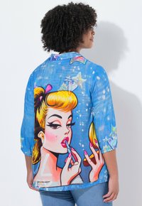 Femme avec des cheveux bouclés portant une chemise bleue sur laquelle est représentée une femme blonde de style cartoon en train d'appliquer du rouge à lèvres tout en tenant un miroir de poche.