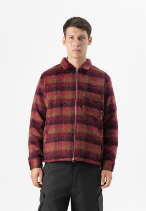 Man wearing a zippered, red and green checkered jacket with a collar, standing against a white background. He has short hair and a neutral expression.