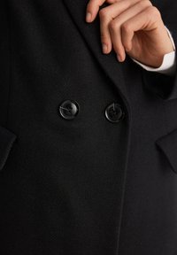 Czarny wełniany płaszcz o teksturowanym wykończeniu, podwójnym zapięciu i błyszczącymi czarnymi guzikami. Widoczna dłoń poprawiająca kołnierz.