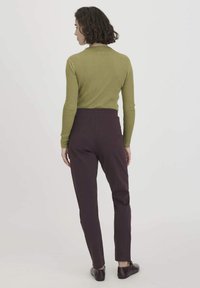 Vrouw met kort gekruld haar, die een olijfgroene geribbelde langemouwtop, donkere paarse broek en zwarte schoenen draagt, staand met haar rug naar de camera gekeerd.