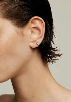 Pendiente de aro pequeño en metal dorado con tres piedras preciosas rosas, con un diseño cilíndrico elegante. Lleva puesto en la oreja.