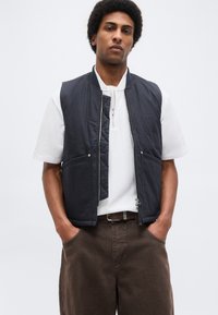 Νεαρός άνδρας που φοράει λευκό κοντομάνικο πουκάμισο, μαύρο γιλέκο χωρίς μανίκια με φερμουάρ και καφέ παντελόνι, με τα χέρια στις τσέπες.