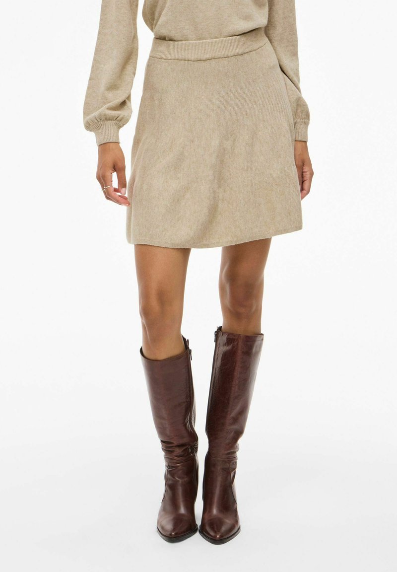 Minigonna beige in maglia con vita aderente, abbinata a stivali in pelle marrone alti fino al ginocchio con zip laterale. Texture liscia, design minimalista.