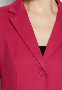 Blazer fucsia in tessuto texturizzato, con revers e chiusura a un bottone sul petto, indossato su una maglia nera.