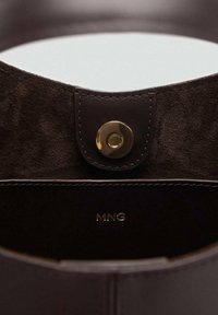 Intérieur en daim brun foncé d'un sac à main avec un miroir circulaire en haut, sécurisé par un bouton-pression doré, comportant le logo embossé "MNG."