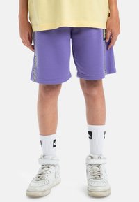Paarse shorts met zijdetails, gecombineerd met witte ankelsokken met zwarte logo's en witte hoge sneakers.