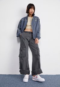 Grå vindjakke med gule detaljer, beige cropped top, grå cargobukser med flere lommer, kombineret med hvide sneakers.