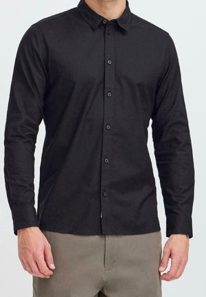 Mann trägt ein schwarzes Hemd mit langen Ärmeln und beige Hosen, steht vor einem einfarbigen hellen Hintergrund.