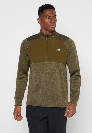 Uomo che indossa un pullover a mezza zip verde oliva e verde scuro con il logo New Balance, in piedi contro uno sfondo chiaro uniforme.