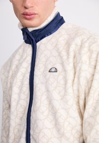 Chaqueta de forro polar en color crema con un patrón en relieve beige. Presenta un cuello con cremallera en navy y un logo en el pecho. Textura suave y diseño casual.
