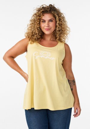 Kobieta o kręconych włosach, ubrana w żółty top bez rękawów z napisem "Summer" i grafiką słońca, stoi z jedną ręką na biodrze, widoczny kwiatowy tatuaż.