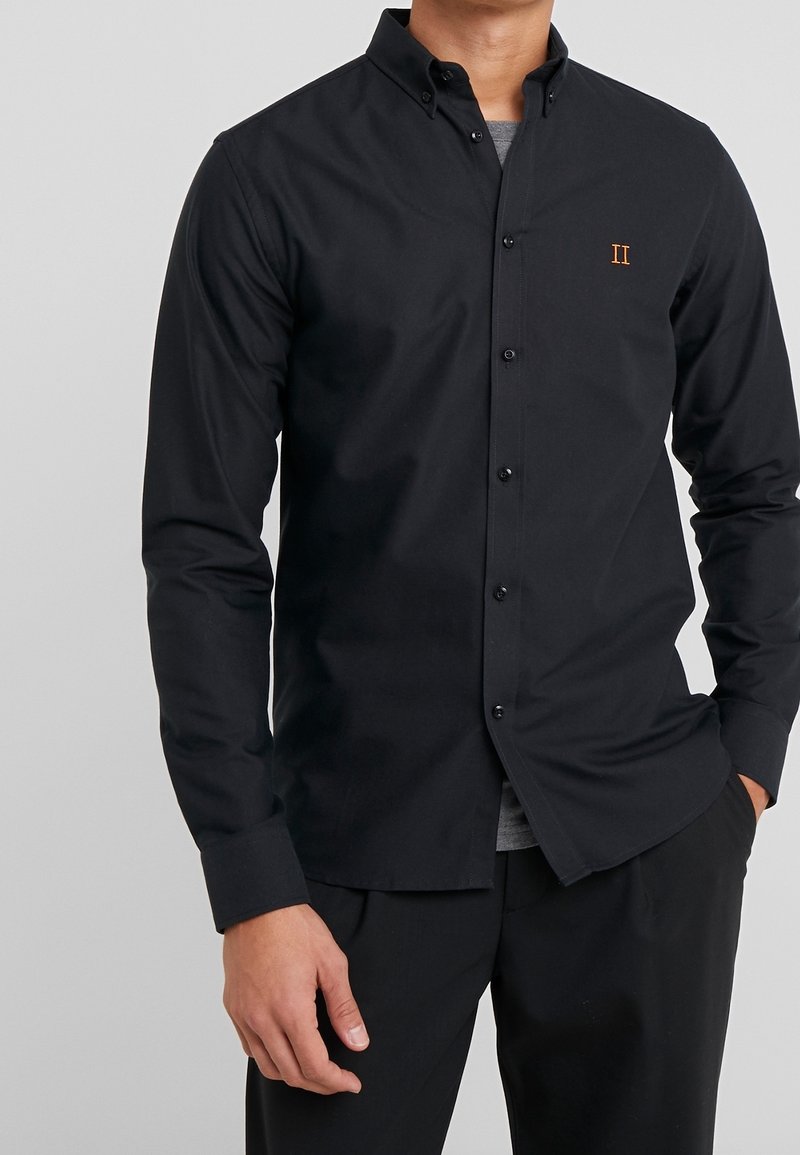 Sort langærmet skjorte med knaplukning på kraven, brystlogo i orange og en lige afskåret kant. Glat stof med en tætsiddende pasform.