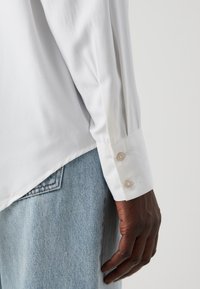 White long-sleeve shirt with a smooth texture, featuring buttoned cuffs. Worn with light blue denim, showing a relaxed fit.