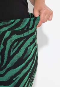 Grön och svart zebra-randig kjol med en texturerad tyg. Modellen håller i midjebandet och visar upp designen och materialet.